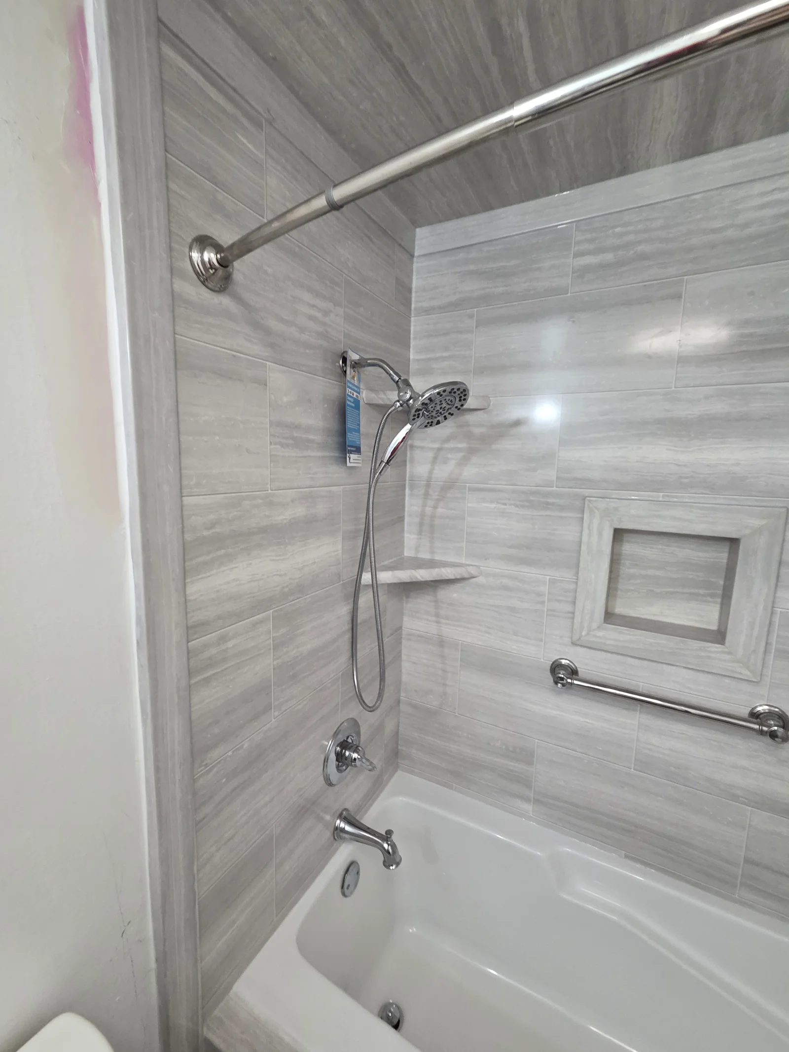 Gray wood-look porcelain tile tub surround with recessed niche, tiled corner shelf, chrome grab bar, and chrome shower fixtures