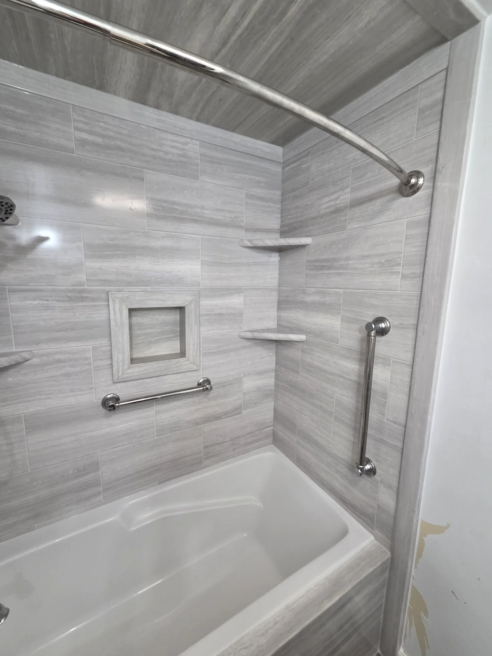 Gray wood-look porcelain tile shower surround with recessed niche, corner shelves, chrome grab bars, and curved shower rod over white tub